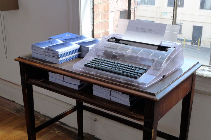 documentation photo of a desk with stacks of booklets and a typewriter on top of it