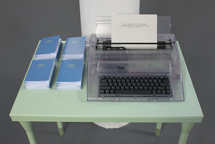 documentation photo of a green metal desk with stacks of booklets and a clear plastic typewriter on top of it and a note in the typewriter