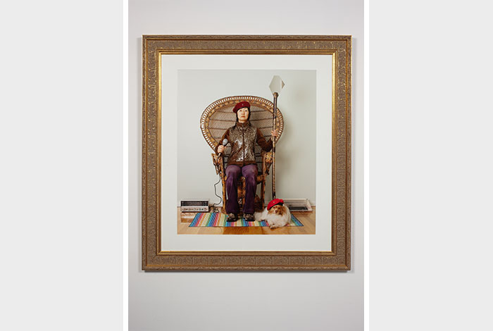 documentation photo of an ornately framed color photograph of Jennifer Moon wearing a beret sitting in a wicker chair holding a microphone in one hand and a staff in the other and a small dog wearing a beret at her feet