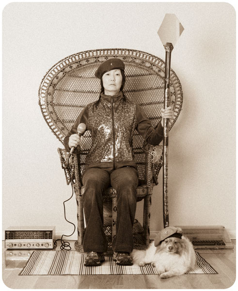 Remake of a famous Huey P. Newton photograph with Jennifer Moon wearing a beret sitting in a wicker chair holding a staff in one hand and a microphone in the other with Mr. Snuggles at her feet also wearing a beret