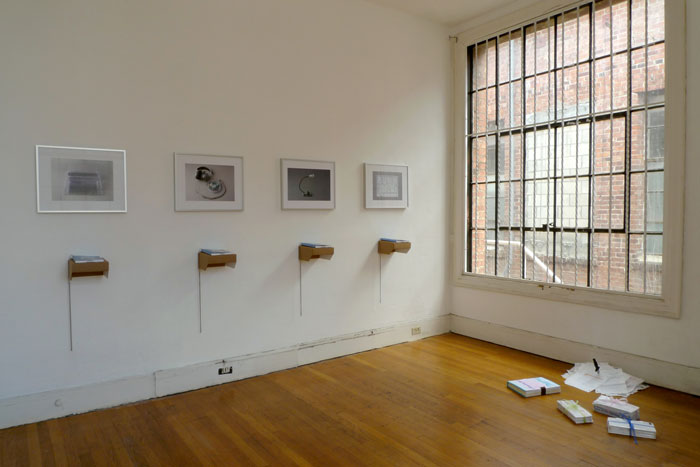 documentation photo of a room containing four framed photographs and four books atop cardboard shelves underneath each framed photo and sculptural objects on the floor