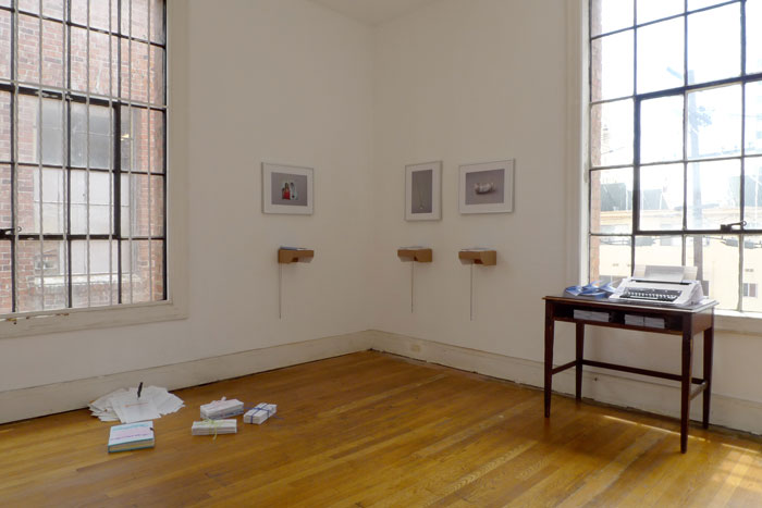 documentation photo of a room containing sculptural objects on the floor, three framed photographs with three books atop cardboard shelves underndeath each photo, and a desk containing stacks of booklets and a typewriter