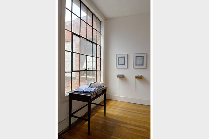 documentation photo of a room containing a desk with stacks of booklets and a typewriter on top of it and two framed photographs with two books atop cardboard shelves underndeath each photo