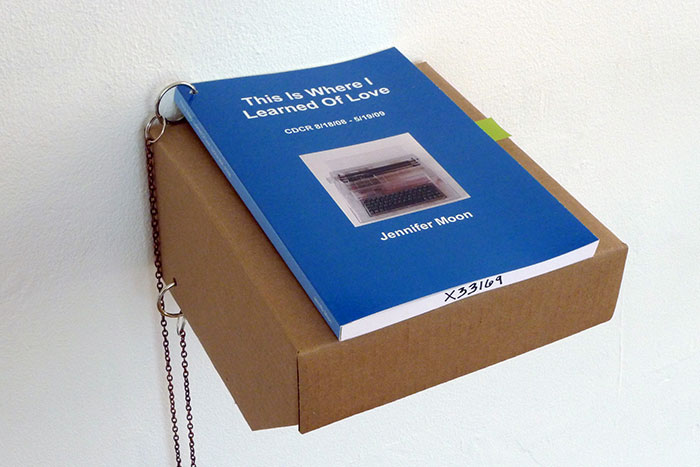 documentation photo of a book on a cardboard shelf with a chain attaching the book to the shelf