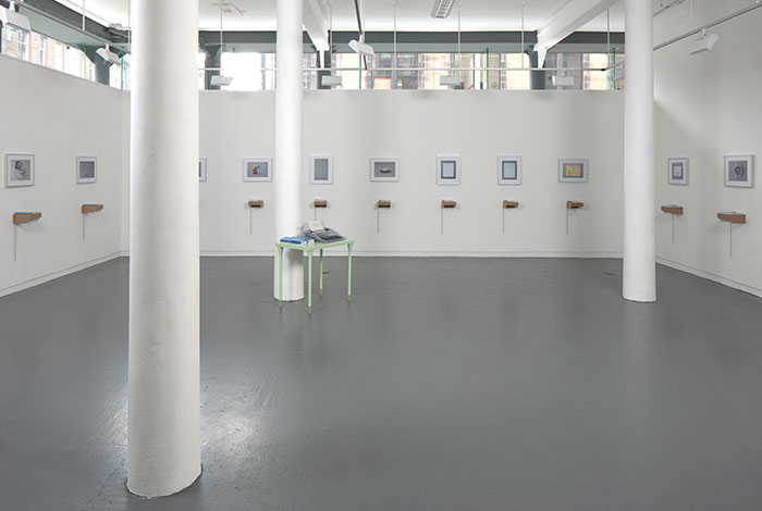 documentation photo of a large room with three pillars containing eleven framed photographs and eleven books atop cardboard shelves underneath each framed photograph and a green metal desk with a clear plastic typewriter and stacks of booklets on top of it