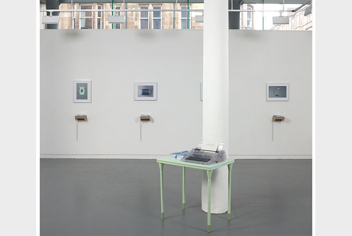 documentation photo of a wall with four hung, framed photographs and four books atop cardboard shelves underneath each framed photograph and a pillar in front of the wall with a green metal desk next to it holding a clear plastic typewriter and stacks of booklets