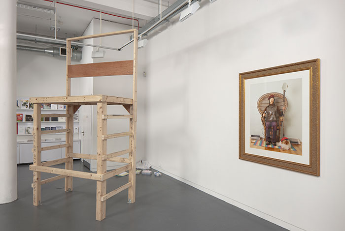 documentation photo of a large wooden platform, sculptural objects on the floor behind the platform, and a large framed photograph hung on a wall